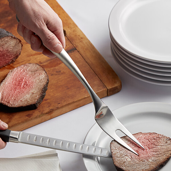 A hand using a Tablecraft stainless steel two-tine fork to cut meat on a plate.