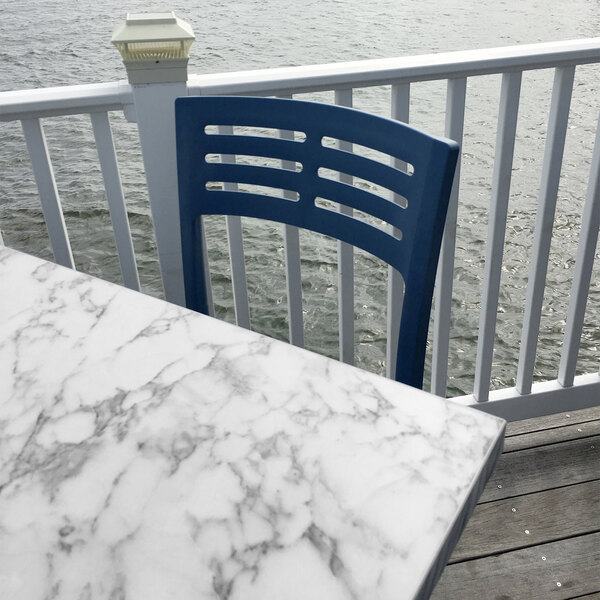 A white Grosfillex marble table top on an outdoor patio with a blue chair and water in the background.