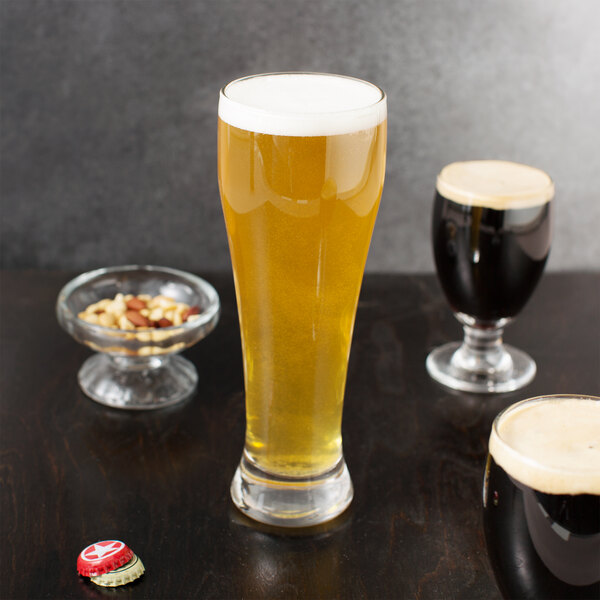 A Libbey giant pilsner glass of beer sits on a table next to a bowl of nuts and a bottle cap.