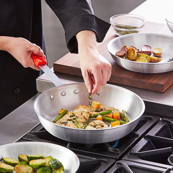 A person cooking food in a Choice aluminum fry pan with a red silicone handle.