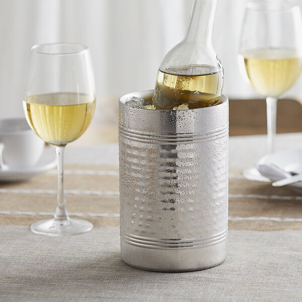 A silver hammered stainless steel wine cooler with a bottle of white wine in it on a table.
