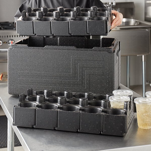 A man using a black Cambro food pan carrier with cup holders to hold a stack of black plastic containers.