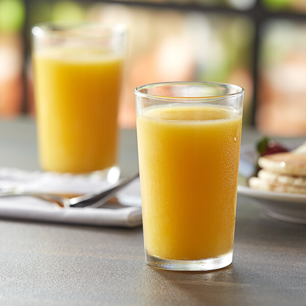 A Duralex glass of orange juice on a table with a plate of pancakes.