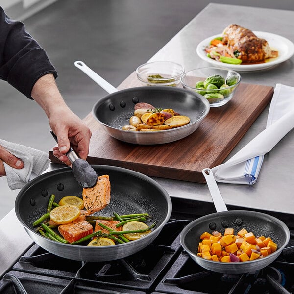A person cooking salmon and vegetables in a Vollrath fry pan.