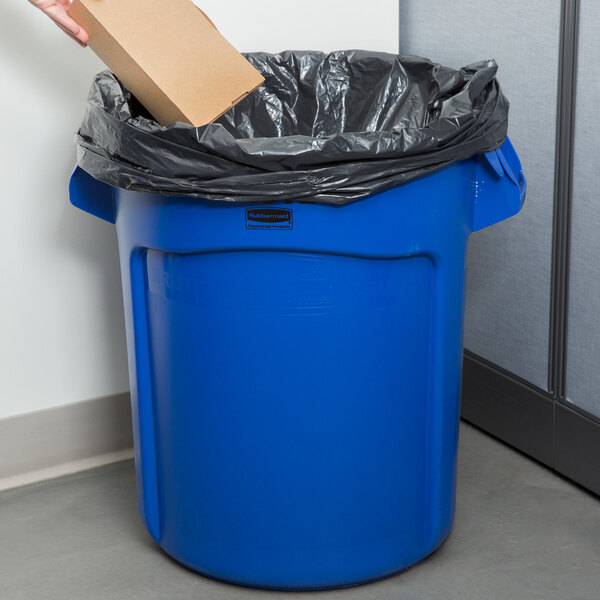 A person putting a brown box into a Rubbermaid blue Brute trash can.