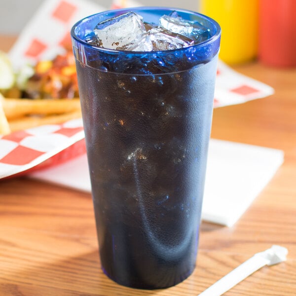 A Carlisle royal blue plastic tumbler on a table with a glass of blue liquid, ice, and a straw.