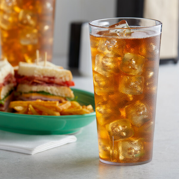 A Carlisle clear plastic tumbler filled with ice tea on a table next to a plate of sandwiches.