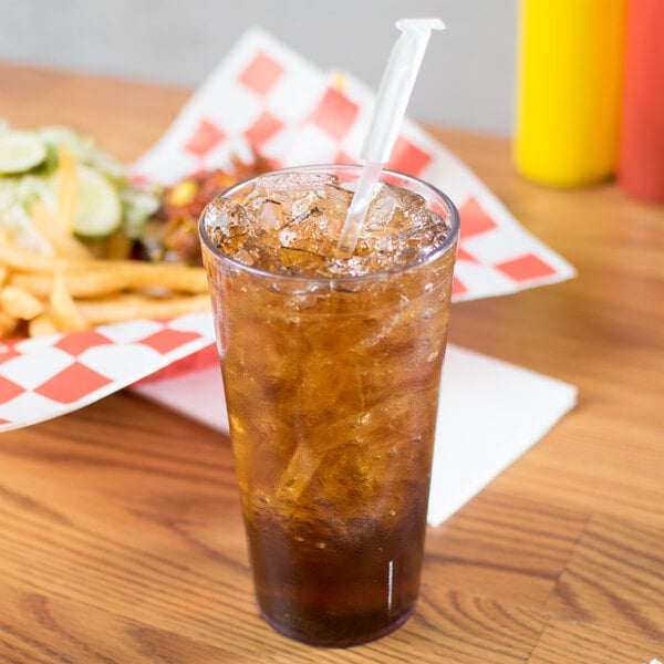 A Carlisle clear plastic tumbler filled with brown liquid and ice on a table with a straw.