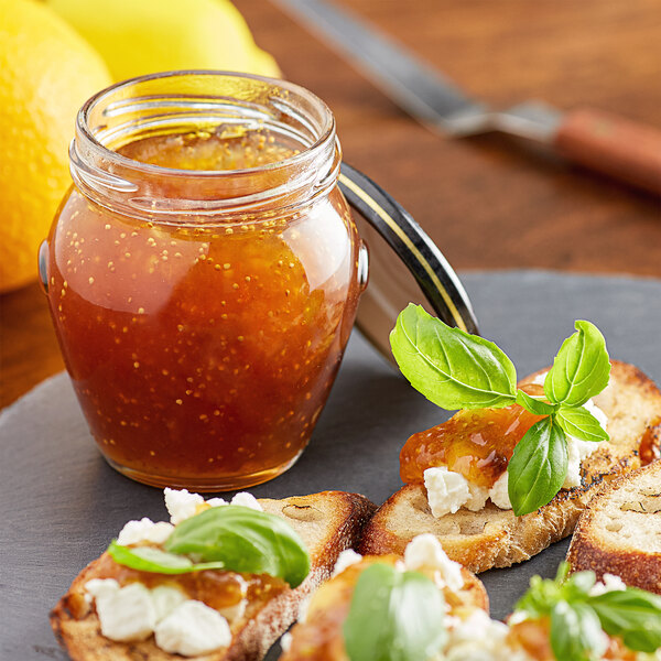 A glass jar of Dalmatia Fig Spread with Fresh Orange next to bread slices with basil leaves.