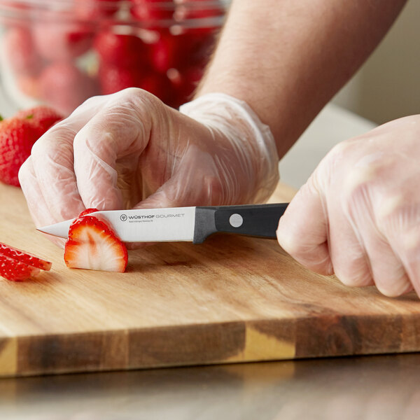 A person in gloves using a Wusthof Gourmet clip point paring knife to cut strawberries on a cutting board.