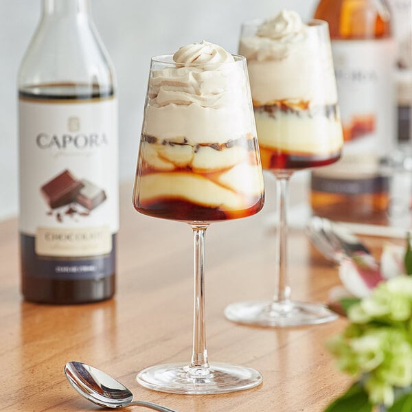 A close-up of a bottle of Capora Chocolate Flavoring Syrup with a glass of brown liquid on a table.