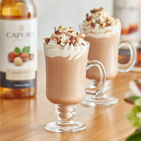 A glass of hot chocolate with whipped cream and nuts next to a bottle of Capora Hazelnut Flavoring Syrup.