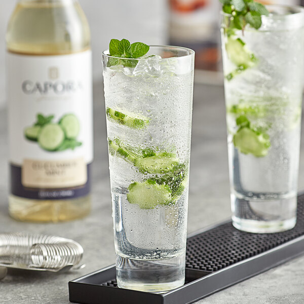 A glass of water with cucumber slices and mint leaves next to a bottle of Capora Cucumber Mint Flavoring Syrup.