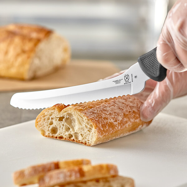 A person using a Mercer Culinary black offset wavy bread knife to cut bread on a cutting board.