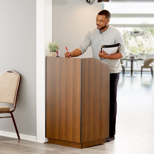 A man standing at a Lancaster Table & Seating walnut host podium writing on a paper.