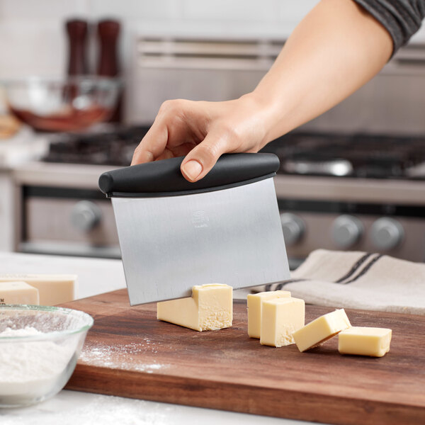 A person using an OXO stainless steel dough cutter with a black handle to cut butter on a cutting board.