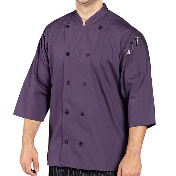 A man wearing a Uncommon Chef eggplant chef coat standing in a professional kitchen.