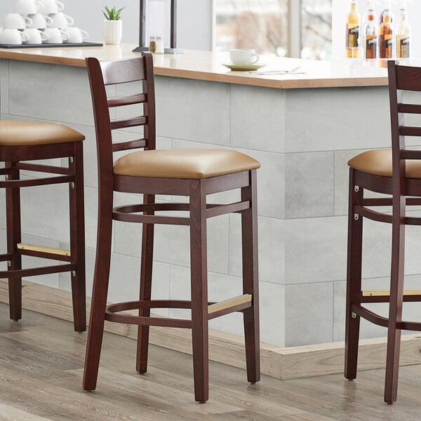 Three Lancaster Table & Seating mahogany wood bar stools with light brown vinyl seats.