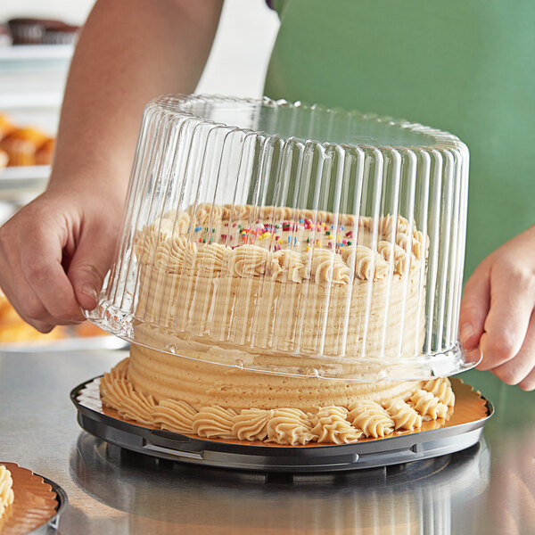 A person holding a Baker's Mark cake with a clear plastic dome over frosting