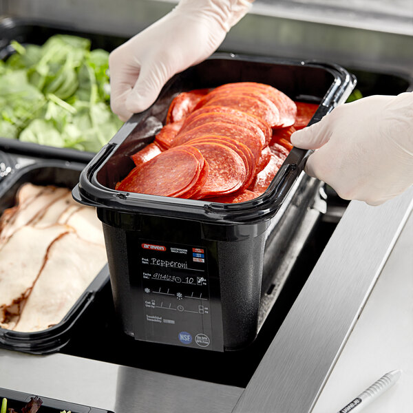 A gloved hand holding an Araven black plastic food pan filled with sliced meat.