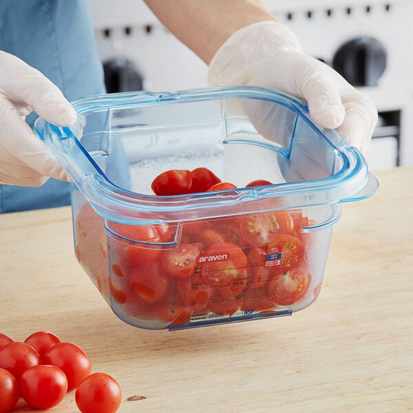 A person in gloves holding an Araven blue plastic food pan with tomatoes inside.