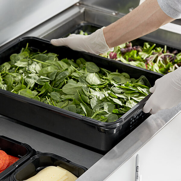 A person wearing gloves putting spinach in an Araven black plastic food pan.