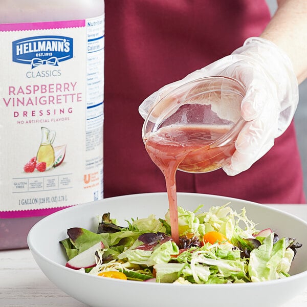 A person pouring Hellmann's raspberry vinaigrette into a bowl of salad.