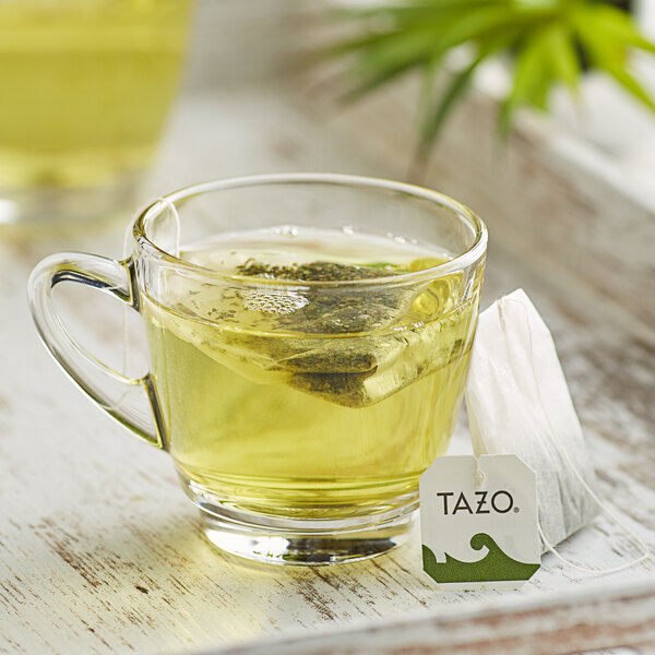 A glass cup of Tazo China Green Tips tea with a tea bag on a tray.