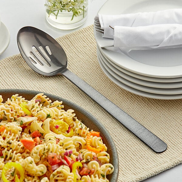 An American Metalcraft hammered black slotted serving spoon in a plate of pasta with vegetables.