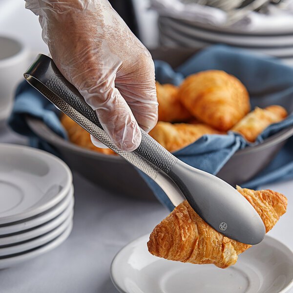 A hand using American Metalcraft hammered black vintage tongs to pick up a croissant.