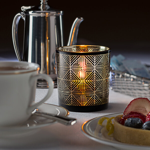 A Hollowick Sussex black and gold geometric glass votive with a lit candle on a table with fruit.