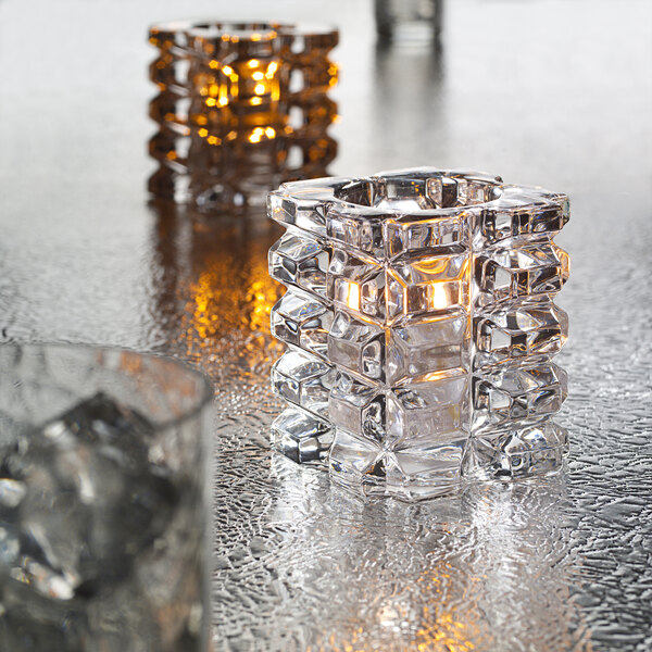 A clear glass Hollowick faceted cube votive with a lit candle inside on a table.