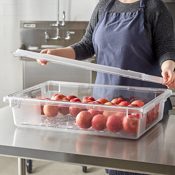 A woman wearing an apron holds a Vigor clear polycarbonate food storage box full of red apples.