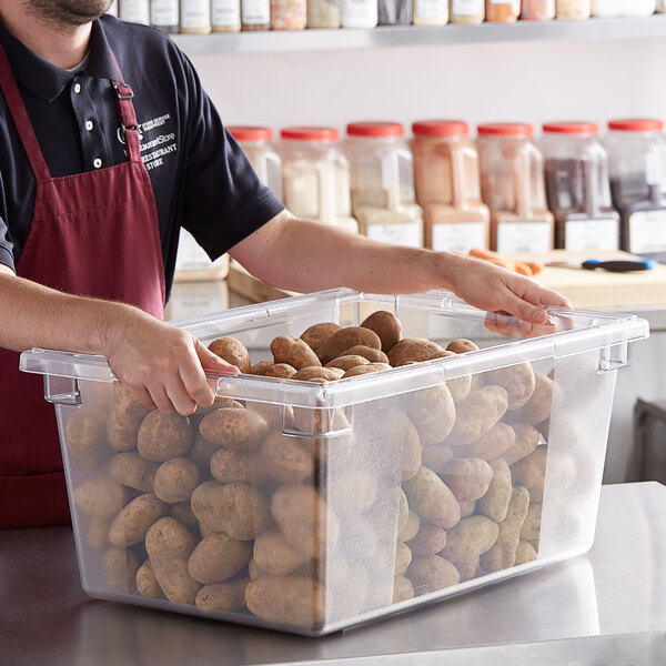 A person in a red apron holding a Vigor clear polycarbonate food storage box filled with potatoes.