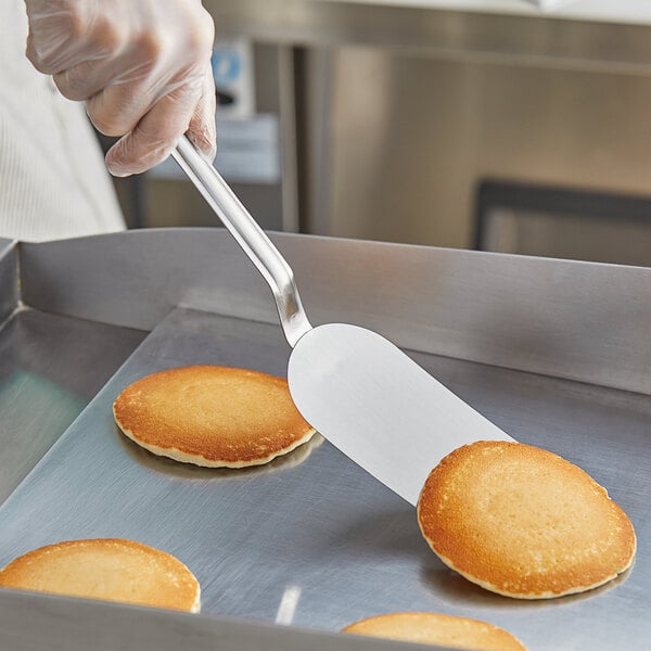 A gloved hand uses a Vollrath stainless steel spatula to lift a pancake from a table.