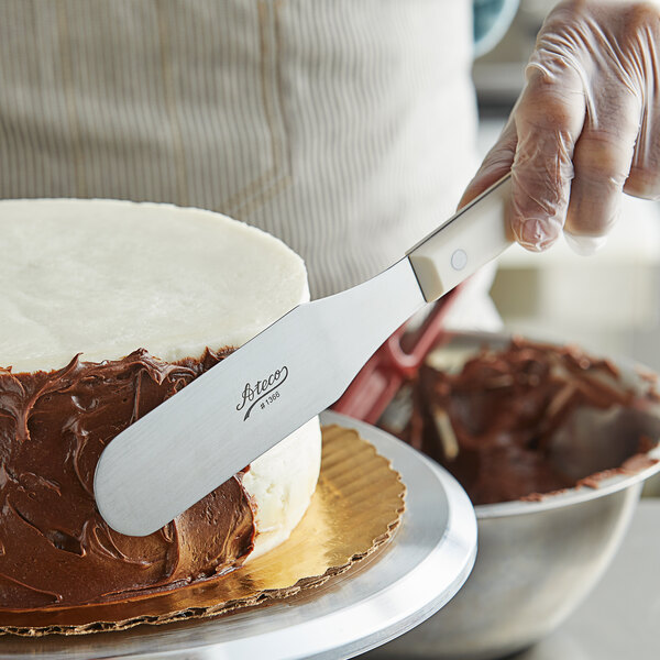 A person using an Ateco straight baking spatula to cut a chocolate frosted cake.