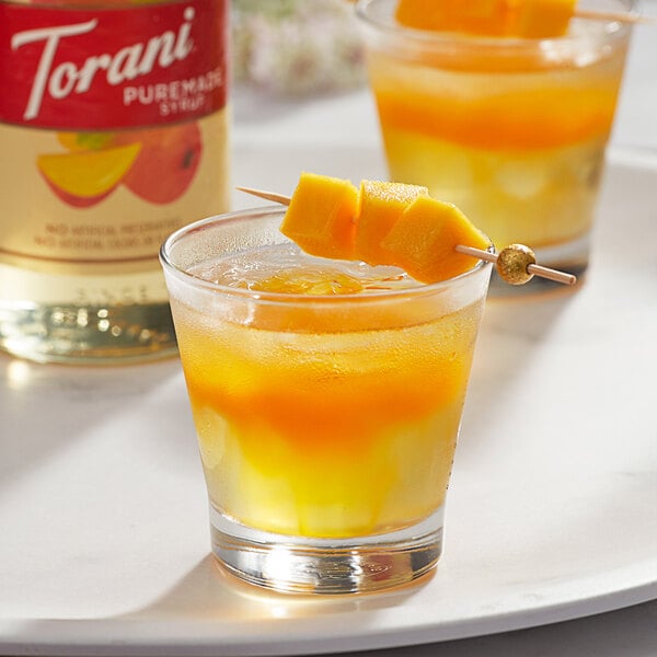 A glass of orange liquid with a slice of mango on a toothpick over a bottle of Torani Mango Flavoring Syrup.