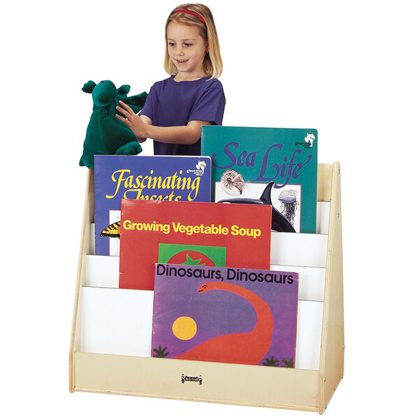A young girl wearing a purple shirt stands next to a Jonti-Craft double-sided book stand filled with children's books.