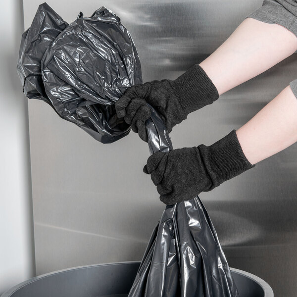 A pair of hands wearing Cordova brown jersey gloves holding a black plastic bag.