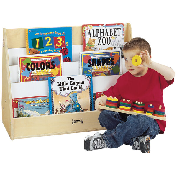 A young boy sitting in front of a Jonti-Craft open back wood book rack holding a toy.