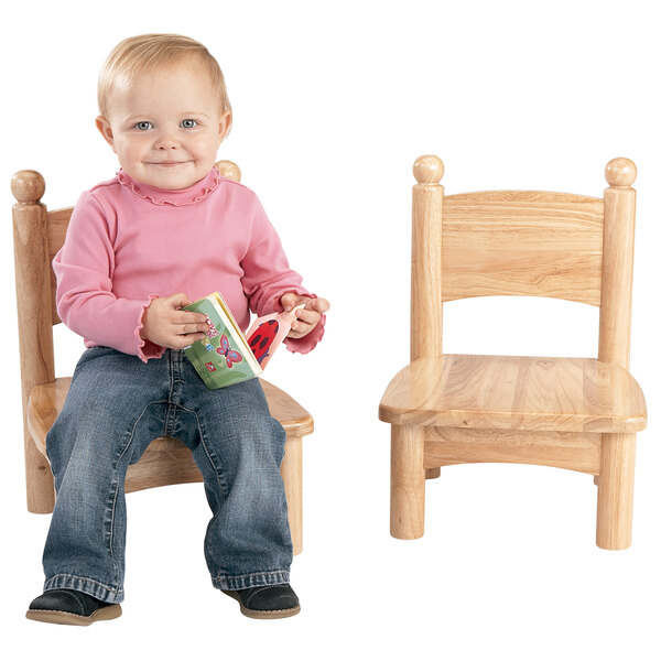 A baby sitting on a Jonti-Craft wooden chair with ball handles holding a book.
