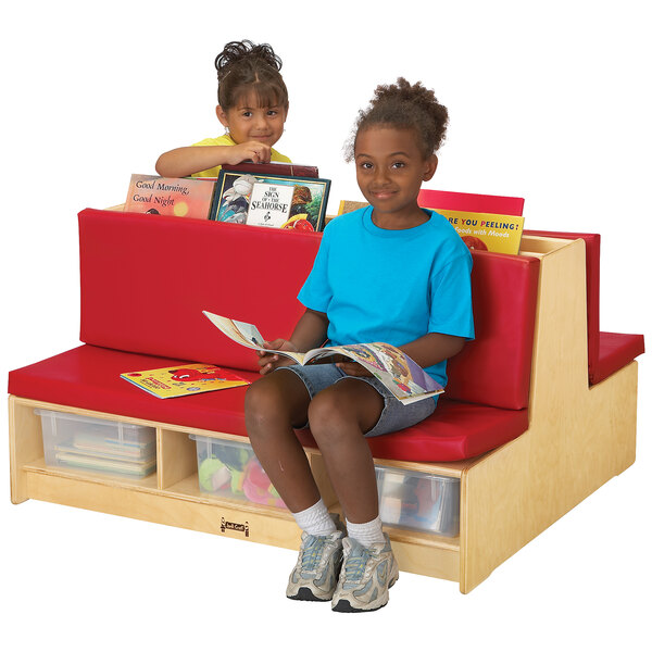 A group of children sitting on a Jonti-Craft double-sided wood bench with red padded seating reading books.