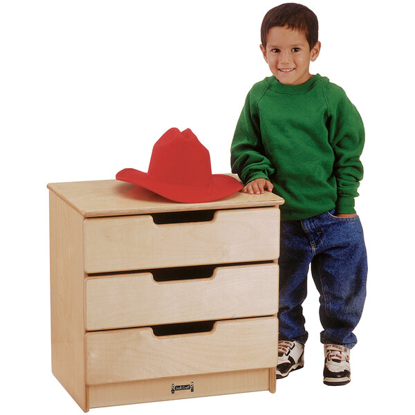 A young boy standing next to a Jonti-Craft wood chest of drawers.