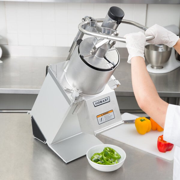 A person using a Hobart food processor to slice green bell peppers on a counter in a professional kitchen.