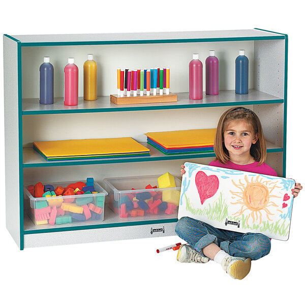 A young girl holding a painting stands next to a teal bookcase with colorful accents.
