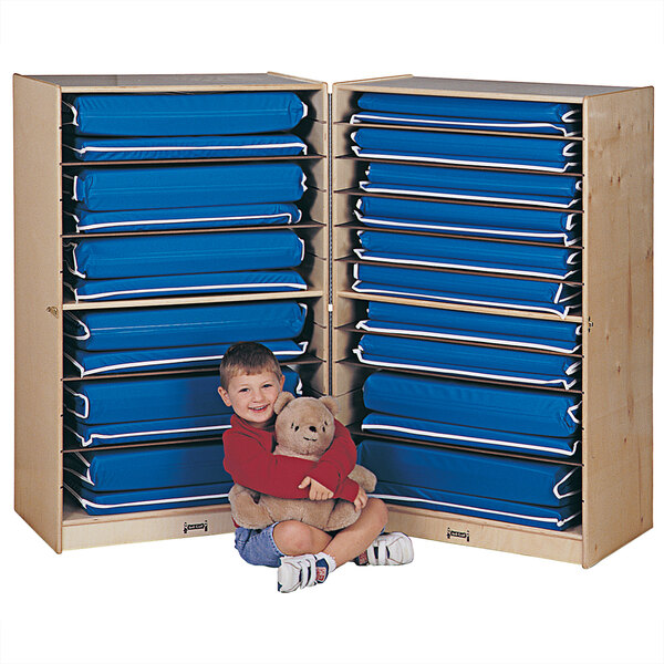 A boy sitting in front of a Jonti-Craft blue storage unit with blue cushions on top holding a teddy bear.