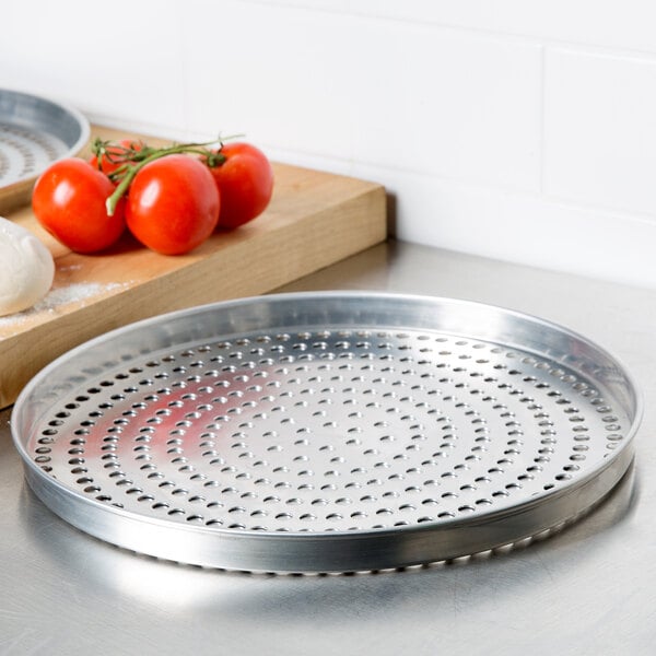 An American Metalcraft standard weight aluminum pizza pan with perforations on a wooden surface with tomatoes and cheese.