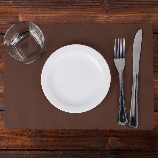A white plate with a glass of water and a fork and knife on a chocolate brown Hoffmaster scalloped paper placemat.