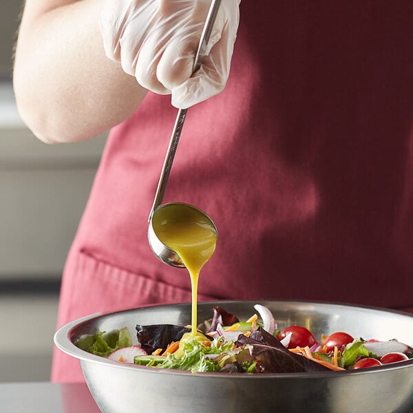 A person using a Vollrath stainless steel ladle to pour yellow dressing into a bowl of salad.