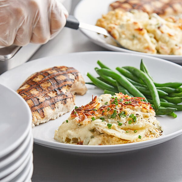 A close-up of a white plate of Spring Glen Fresh Foods white cheddar scalloped potatoes with a person holding a knife.